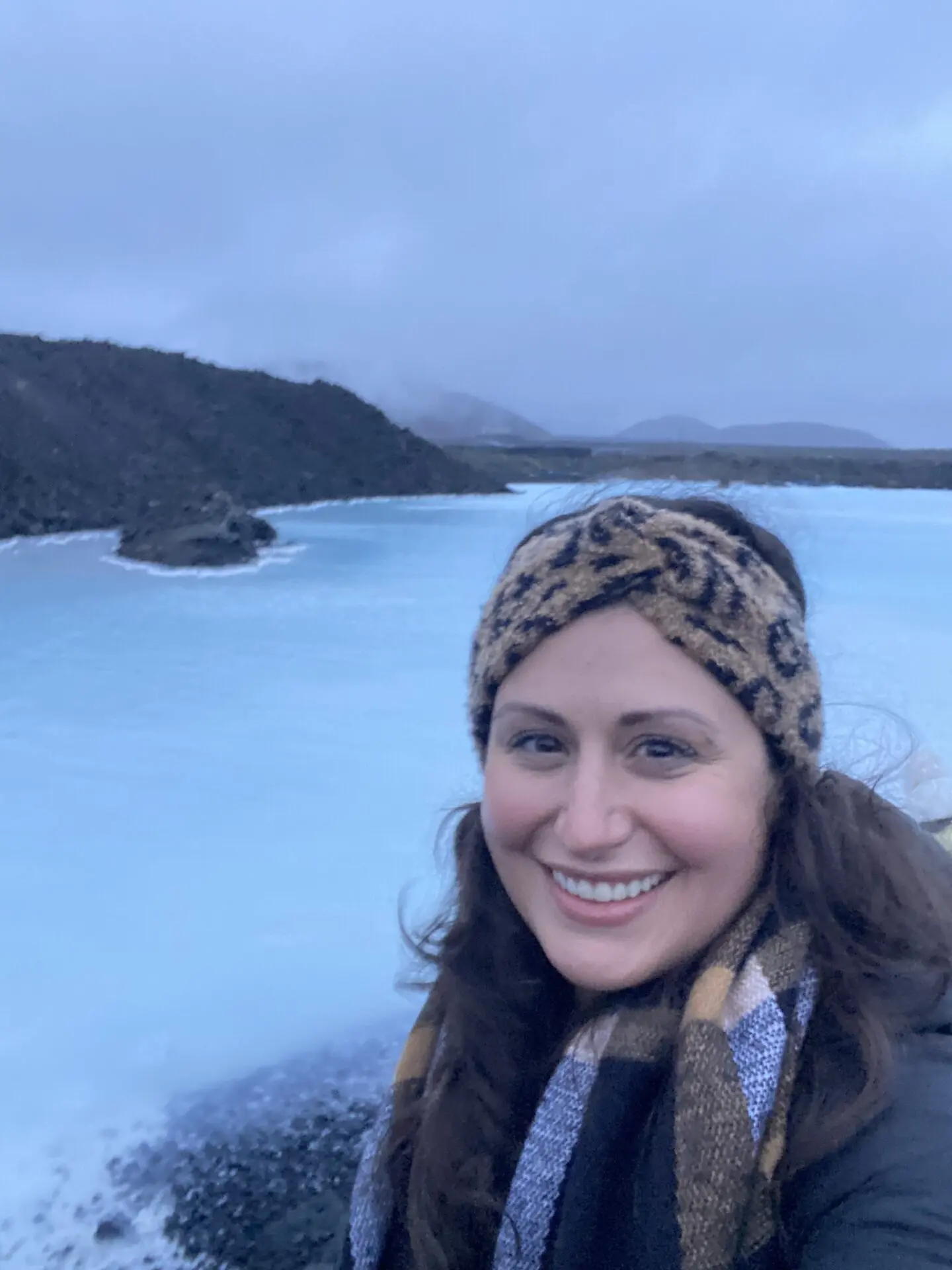 A woman in a scarf standing on the shore of a lake.